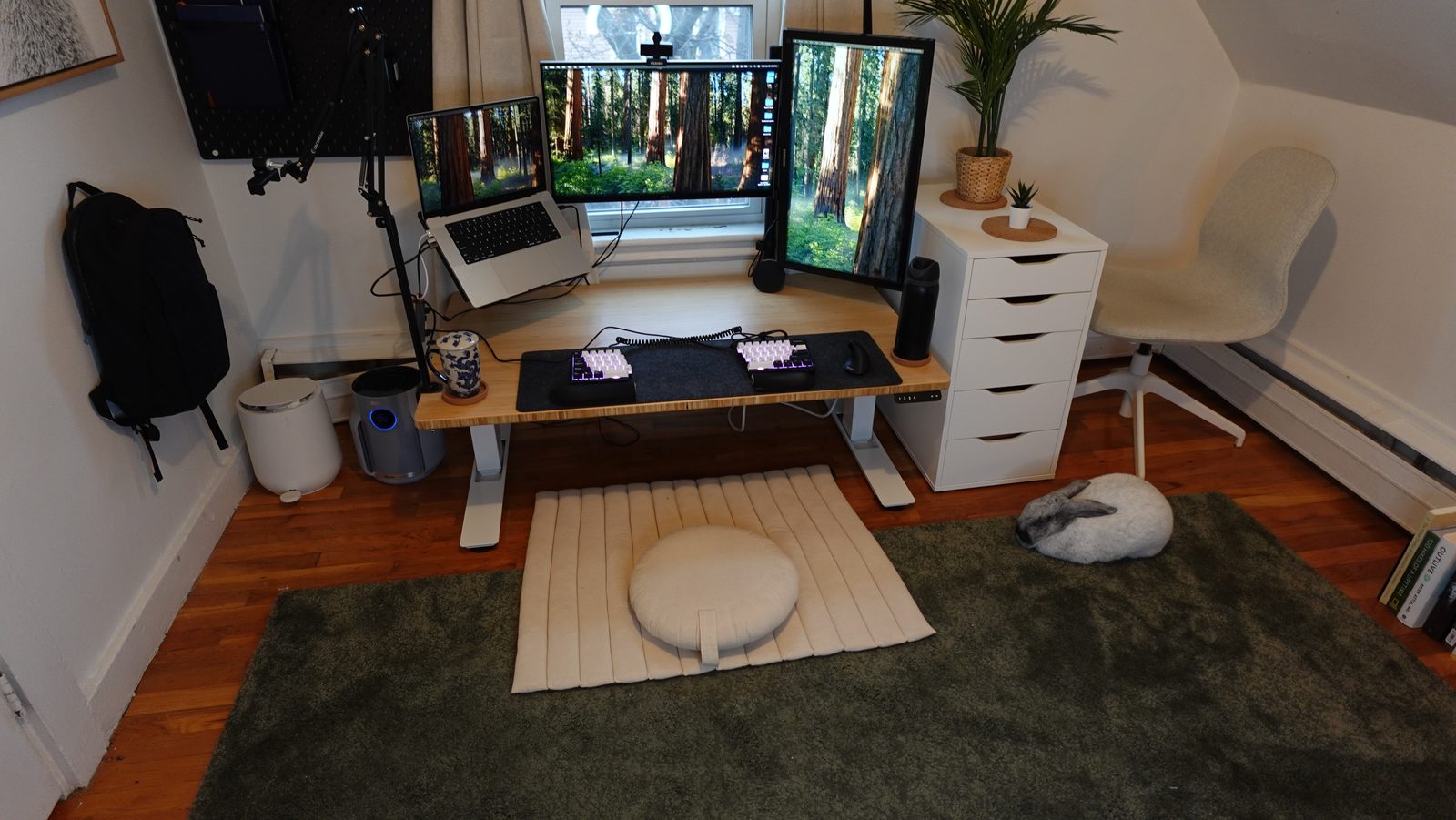 The Grounded floor desk setup — cushion and quilted mat on an olive rug, warm wood floors, plants, and a resting rabbit.